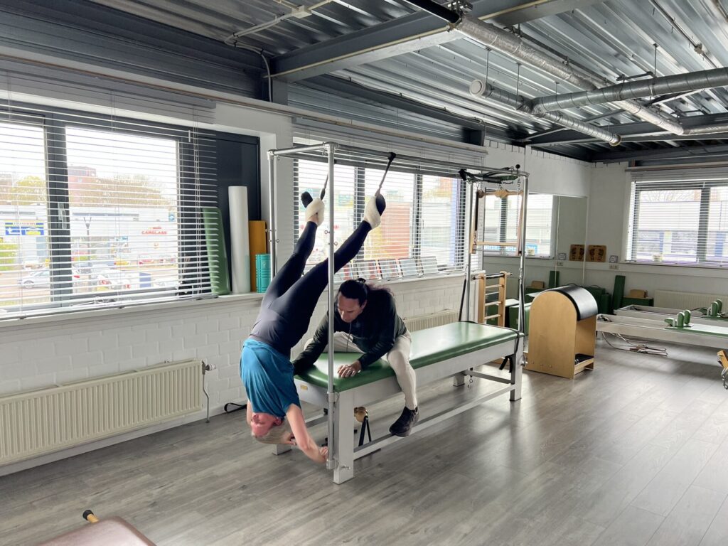 Een pilatesleerling hangt ondersteboven aan de fuzzy straps van de Cadillac terwijl een trainer haar nauwkeurig begeleidt. De oefening wordt uitgevoerd in een lichte, professionele pilatesstudio met grote ramen en een houten vloer.
