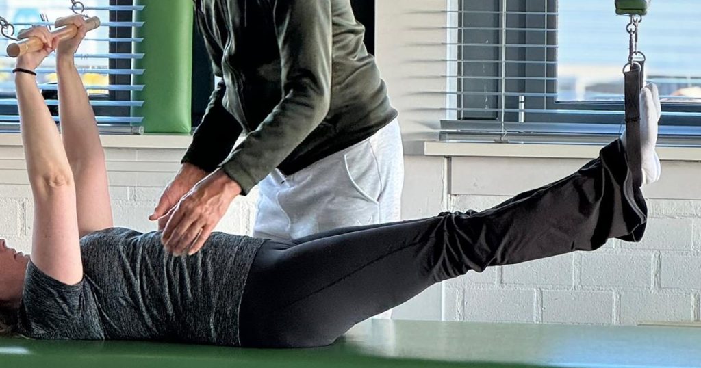 Persoon op reformer, handen van trainer zichtbaar, geeft instructie: "Adem in... en uit... trek je buik op."